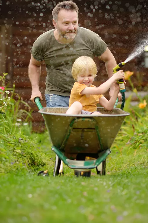A Father and Son Spray a Garden Hose During a Wheelbarrow Ride