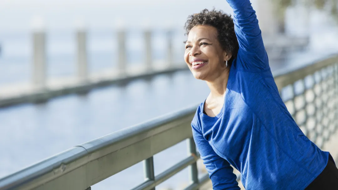 A woman is exercising outdoors, stretching her arm over her head, near the water.