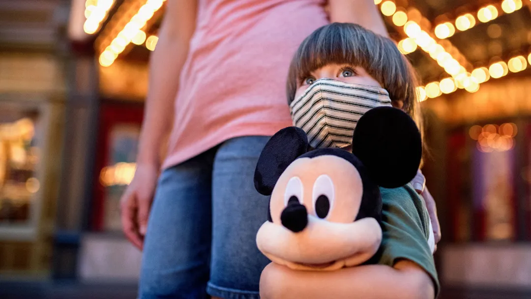Little boy at Disney in a mask holding a stuffed Mickey Mouse.