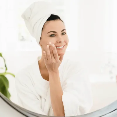 Woman in white bathrobe and towel smiling in mirror while applying skincare cream to her face in a bright bathroom