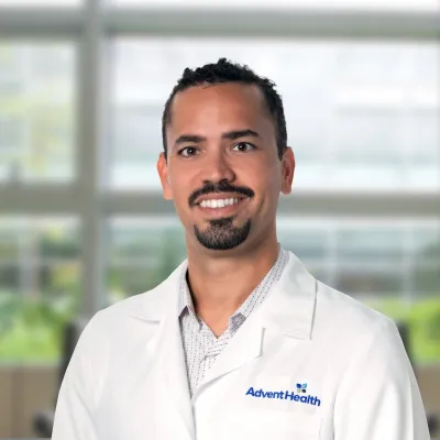 Dr. Paoli, a tan-skinned man with dark hair and facial hair, smiles at the camera. Dr. Paoli is wearing a white medical coat, and he stands in front of a blurred, office-style background.