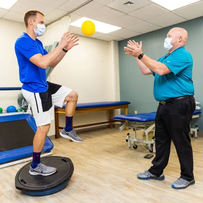 A patient balancing on a medicine ball while catching a small ball from his instructor