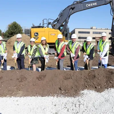Dignitaries wearing hard hats use shovels to dig into pile of dirt in front of construction equipment during groundbreaking ceremony.