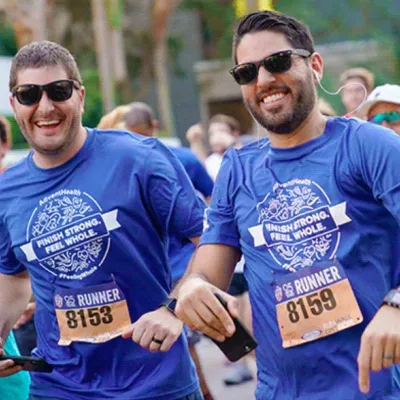 Two men smile at the camera while running Track Shack's annual Corporate 5k in downtown Orlando. Their t-shirts say, "Finish Strong, Feel Whole."
