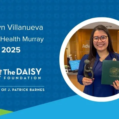 Jocelyn Villanueva, a woman with dark hair wearing navy blue scrubs, holds her DAISY Award in front of a reception area