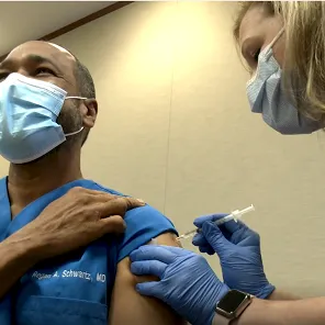 Dr. Schwartz rolls up his blue scrub sleeve as nurse administers the COVID-19 Vaccine