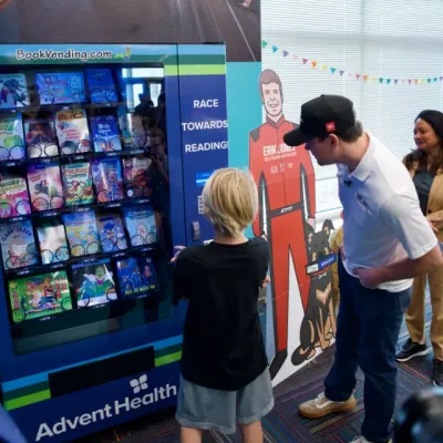 Erik Jones, driver of the No. 43 AdventHealth Toyota Camry XSE, helps a Chisholm Elementary School student use the new book vending machine for the first time.