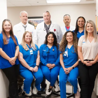 A smiling group of people, some in blue scrubs, and some in white medical coats, stand in a doctor's office. 