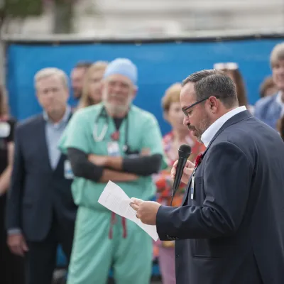 Isaac Sendros, a white man with dark hair, wearing glasses and a grey suit, speaks in front of a group of AdventHealth team members