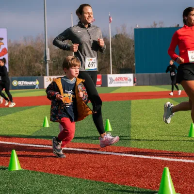People are running on a baseball field. There is one small child in the foreground, and several adults behind him running.