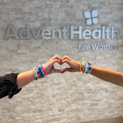 Two hands with friendship bracelets on the wrists form a heart in front of the AdventHealth for Women sign.