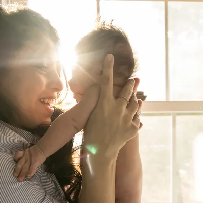 A woman stands in front of a window, holding up a newborn baby and smiling as she looks in his eyes.