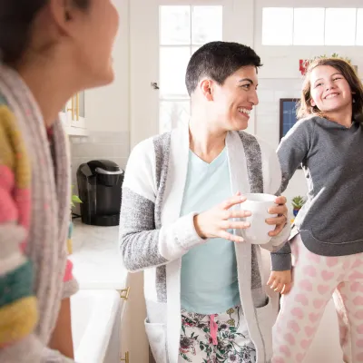 A woman dancing in the kitchen with her daughters and drinking coffee.