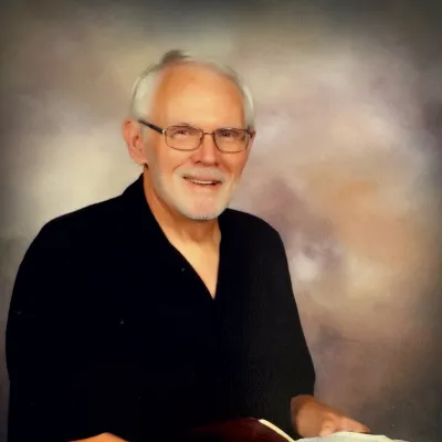 Charlie Williams sits with his Bible open in his hands in front of a beige background. He is a white man with gray hair and a gray beard. He is wearing a black shirt and khaki pants. 