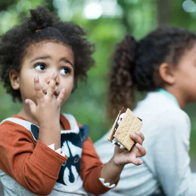 Kids eating s'mores while seating around the campfire in the forest.