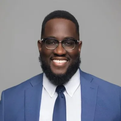 Mack Vendome, an African-American man wearing a blue suit, white shirt, and glasses, smiles in front of a plain gray background.