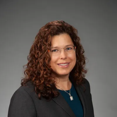 Marcy Cent, the new director of oncology services for AdventHealth Georgia, smiles at the camera in front of a grey backdrop. Marcy is a white woman with curly auburn hair. She is wearing glasses.