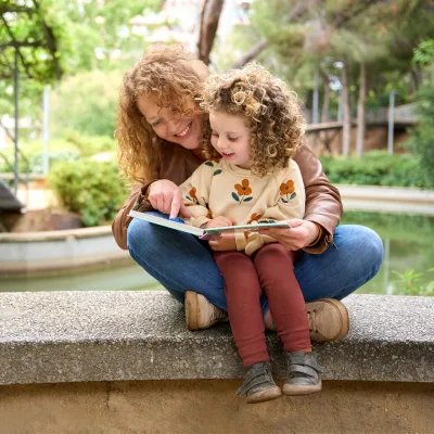 Mother and child reading a book together while sitting on a wall by a river.
