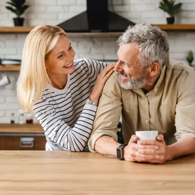 An older couple smiling at each other while in a kitchen.