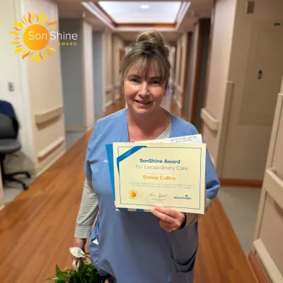 Donna Collins, a white woman in light blue scrubs, smiles holding her SonShine Award certificate.
