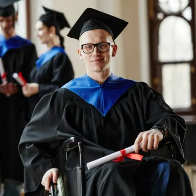 A student in a wheelchair wears a graduation cap and gown and holds a diploma.