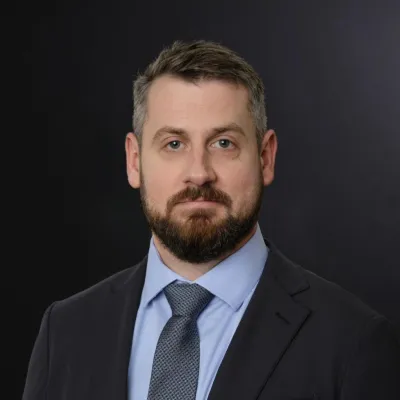 Dr. Nicholas Wells, a white, bearded brunette man, is pictured with a dark gray background. He is wearing a dark colored suit and tie with a blue collared shirt.