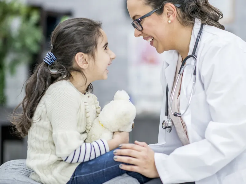 A child sitting on an exam table with her doctor, who has her hand on her knee and is smiling at her.