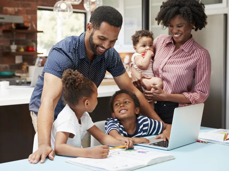 A family of five is in the kitchen, helping two of their children with their homework.