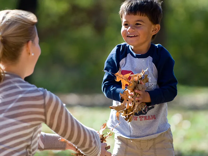 A boy plays happily in the fallen leaves with his mother.