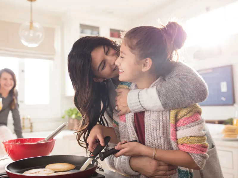 A family of three women are in the kitchen, one daughter hugging the youngest while she flips pancakes.