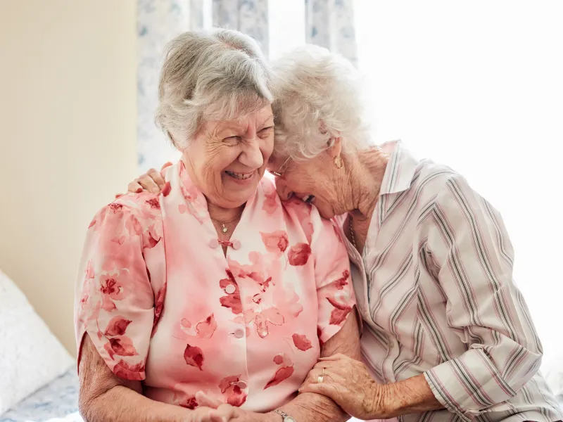 Two older women laugh together while sitting on a bed.