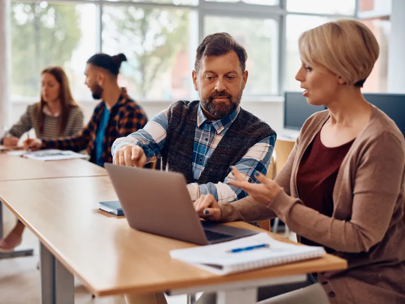 Adult students talking while using laptop and studying together in the classroom.