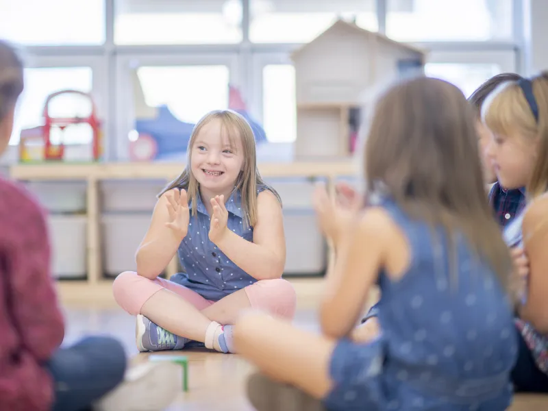 A cute little girl with Down syndrome smiles as she claps along to music with her friends.