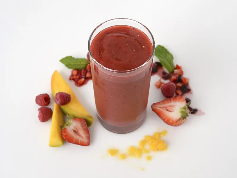 a tall glass of smoothie, surrounded by chopped mango and mixed berries