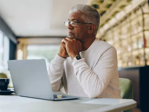 A man sitting at a table in a cafe staring in the distance while his laptop, a cup of coffee and a cup of water sit on the table.