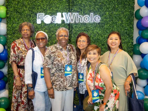Women attending the Women’s Health and Cancer Prevention Event, taking a group picture in front of a green backdrop with a sign that states 'Feel Whole'.