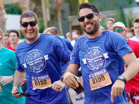 Two men smile at the camera while running Track Shack's annual Corporate 5k in downtown Orlando. Their t-shirts say, "Finish Strong, Feel Whole."