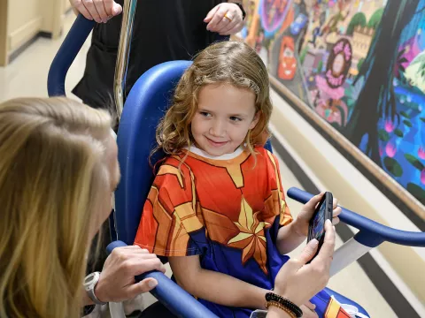Little girl in a wheelchair at AdventHealth for Children wearing a Captain Marvel gown and using the Disney Team of Heroes mobile app.