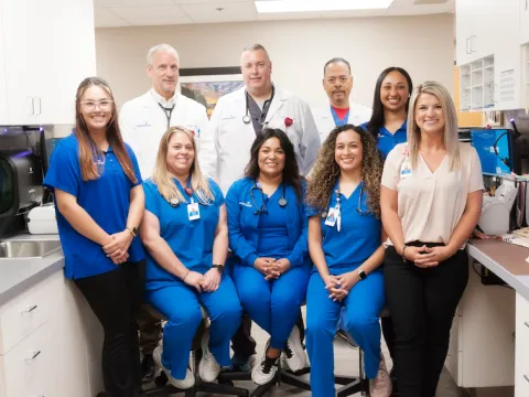 A smiling group of people, some in blue scrubs, and some in white medical coats, stand in a doctor's office. 