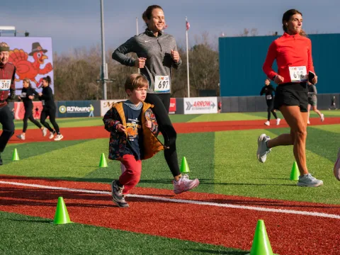 People are running on a baseball field. There is one small child in the foreground, and several adults behind him running.