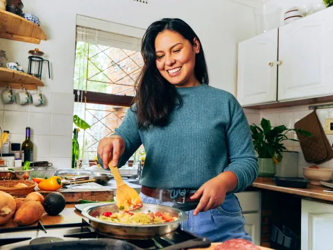 Smiling person stirs vegetables in a pan in a bright, cluttered kitchen.