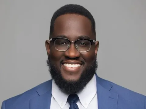 Mack Vendome, an African-American man wearing a blue suit, white shirt, and glasses, smiles in front of a plain gray background.