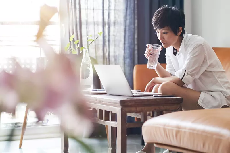 A woman drinks a cup of water while working on her laptop at home