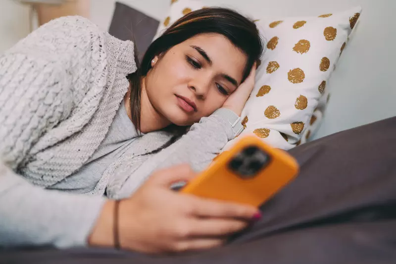 A Woman Lays on a Bed Looking at Her Cell Phone.