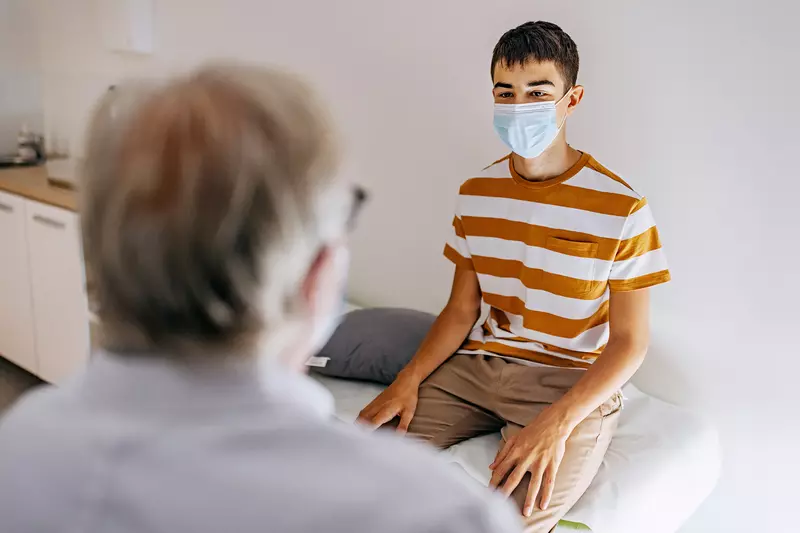 A Teenager Wearing a Face Mask Speaks to His Physician.