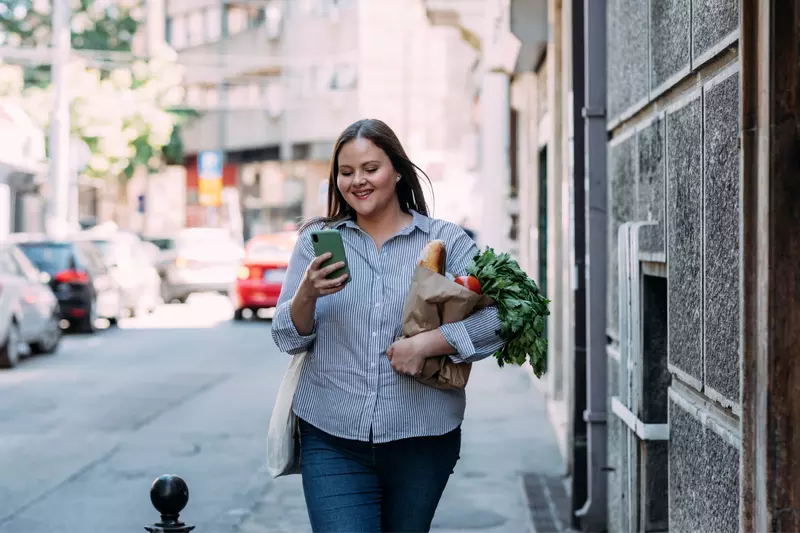 A Woman Walks Down a City Street Reading Her Cell Phone While Carrying Her Groceries  