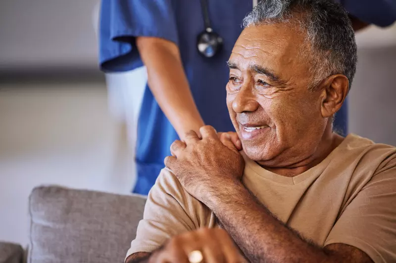 A Senior Man Smiles as a Nurse Puts Her Hand on His Shoulder for Comfort.