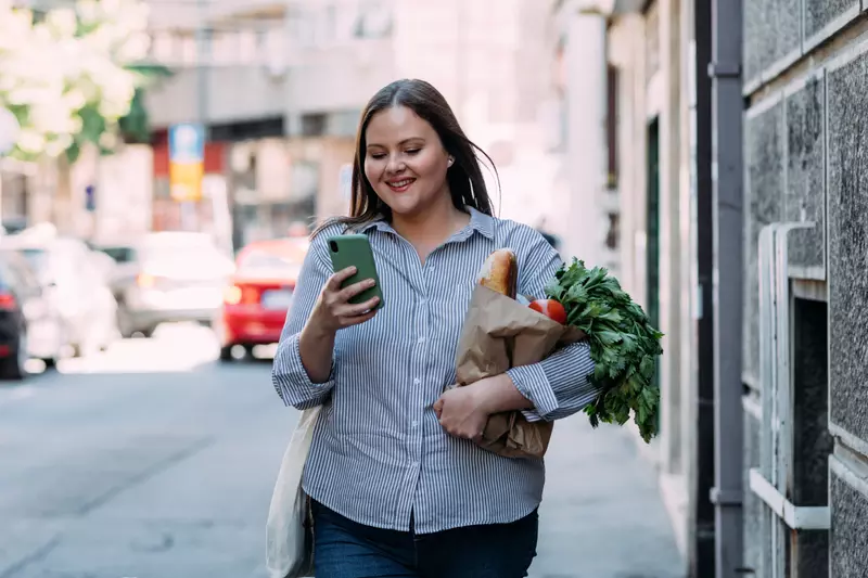 A Woman Walks Down the Sidewalk Checking Her Phone While Carrying Groceries