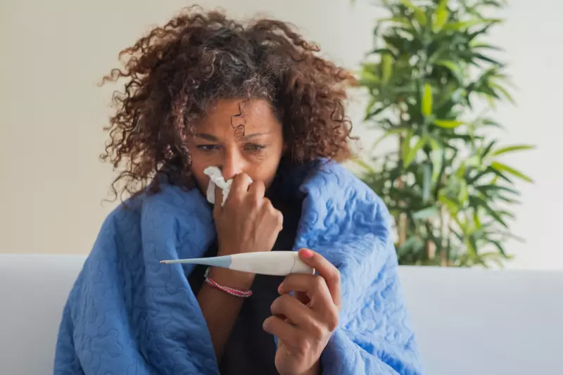 A Woman Blows Her Nose While Sitting on Her Couch at Home Reading a Thermometer 