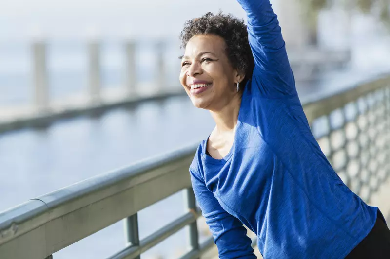 A woman is exercising outdoors, stretching her arm over her head, near the water.